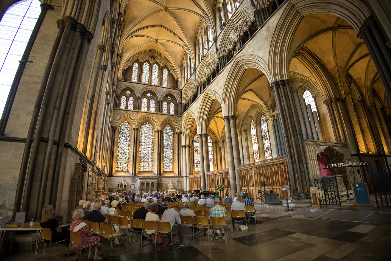 An interior shot of the Cathedral, lit up with warm light. An audience is sat in the foreground, listening to the concert.