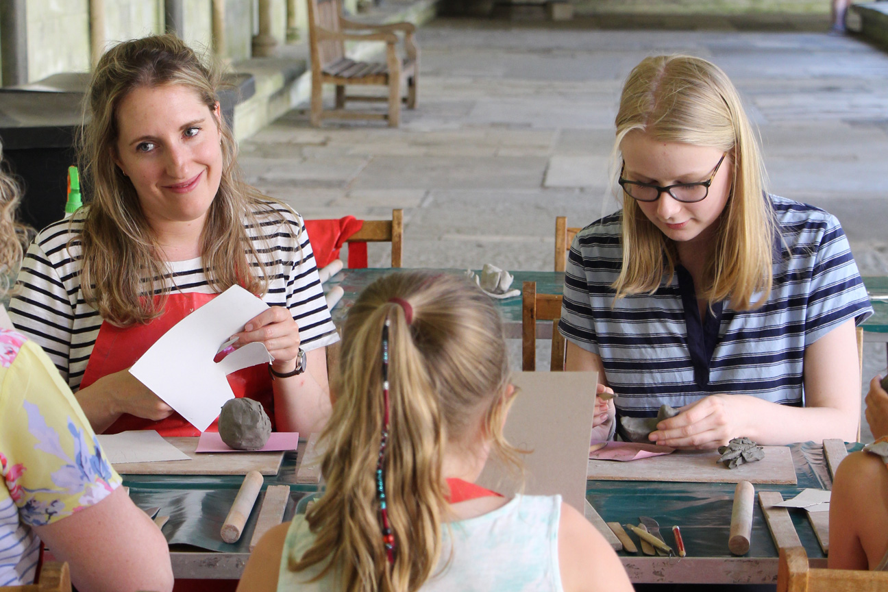 Two adult women sit at a crafts table with a child in the Cloisters of the Cathedral. They are working to make small clay sculptures.