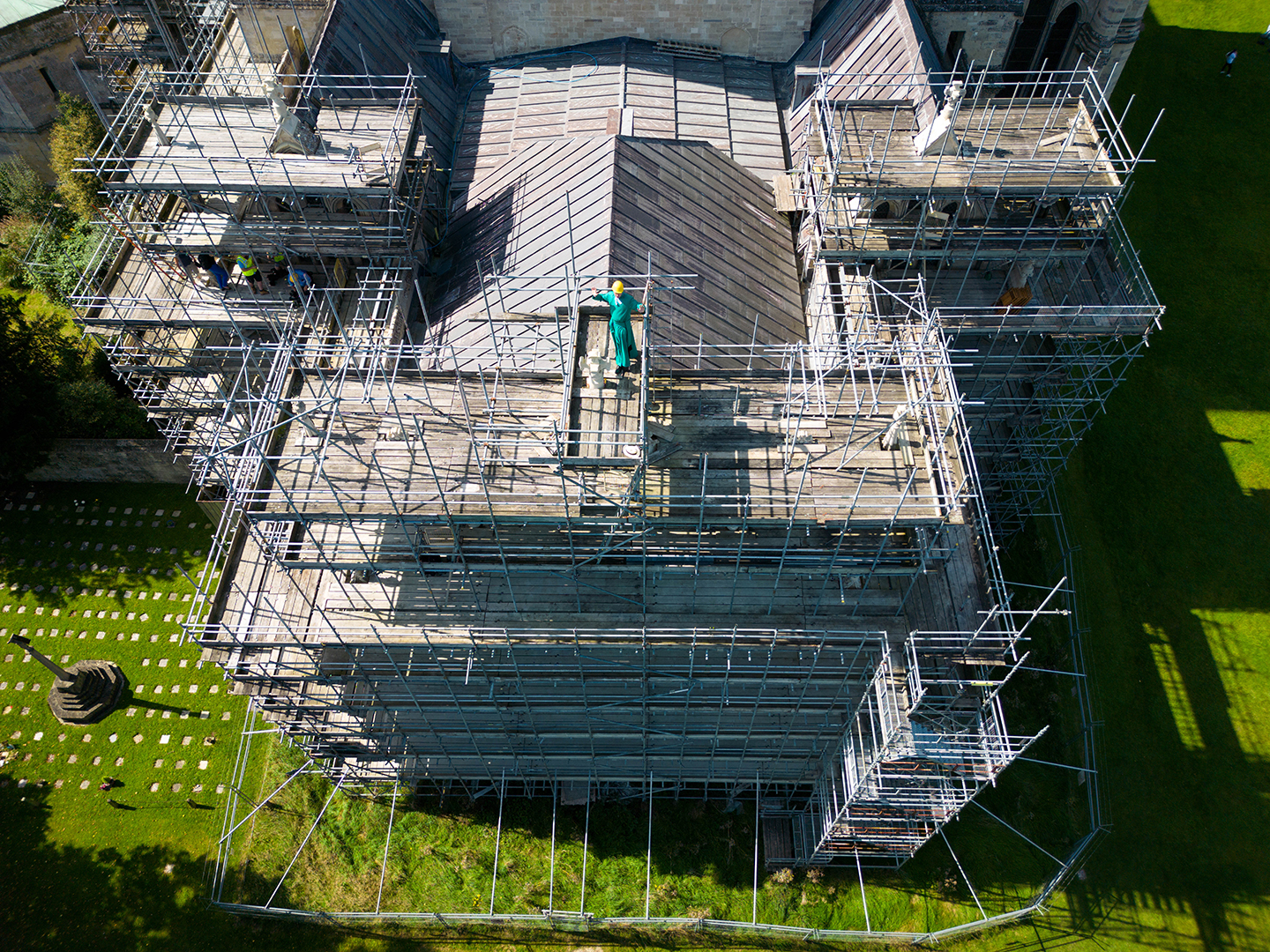An aerial drone shot of Salisbury Cathedral and the scaffolding on the East End. Canon Padley stands atop the scaffolding, next to the final cross, hands raised to the sky.
