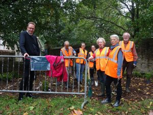 Volunteers in high viz jackets stand in a line, partially behind a metal fence. Canon Treasurer Kenneth Padley is stood to the left of the volunteers. 