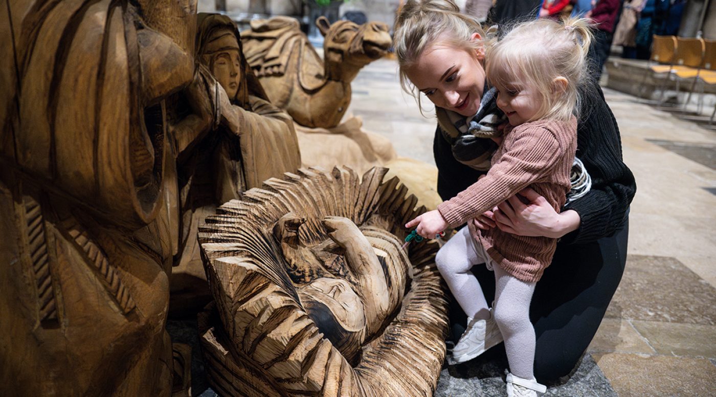 Small child and carer interact with wooden nativity scene